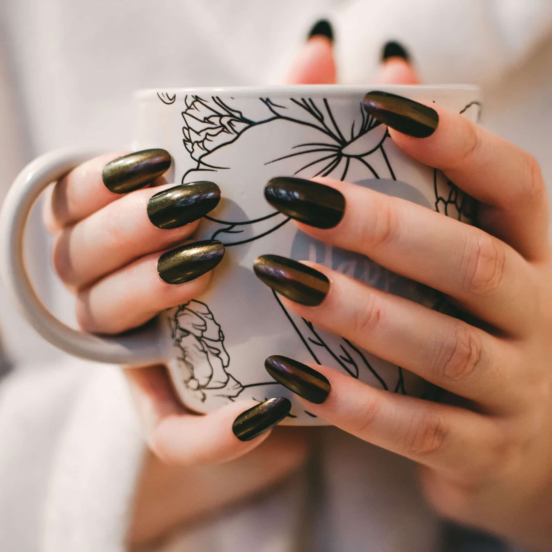 Mulher com unhas pintadas de preto segurando uma caneca branca com detalhes escuros