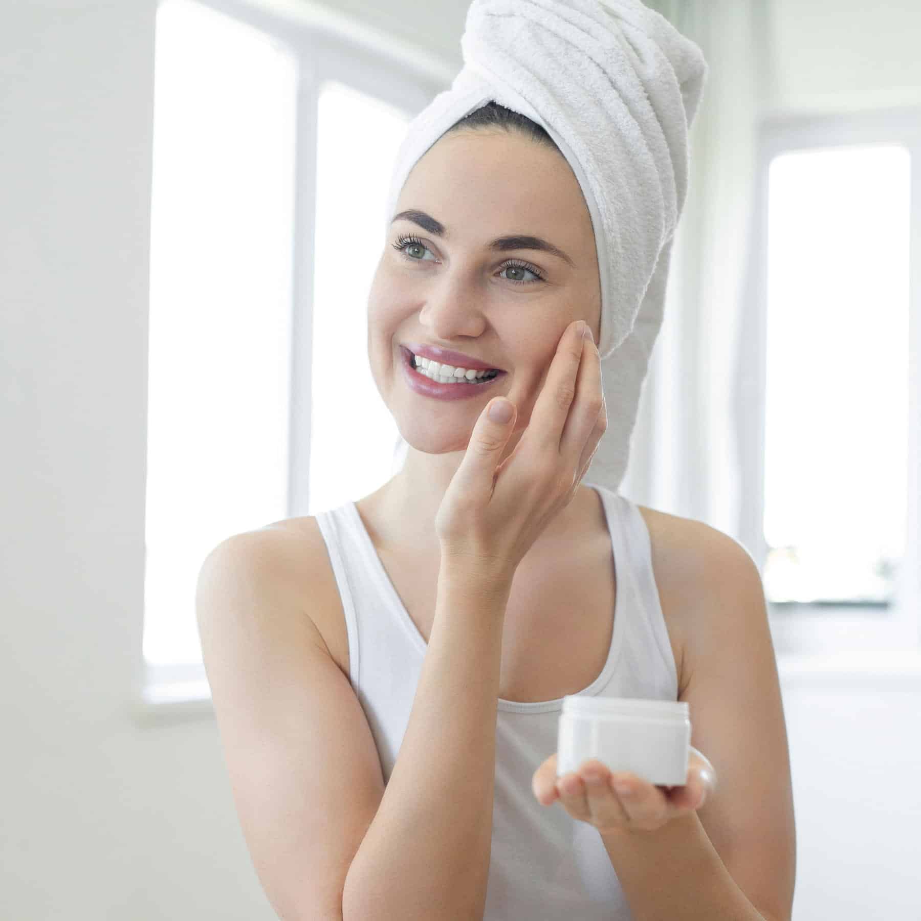 Mulher vestida com regata branca e toalha na cabeça aplicando creme no rosto