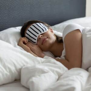 Mulher dormindo com tapa olhos em uma cama com lençóis brancos